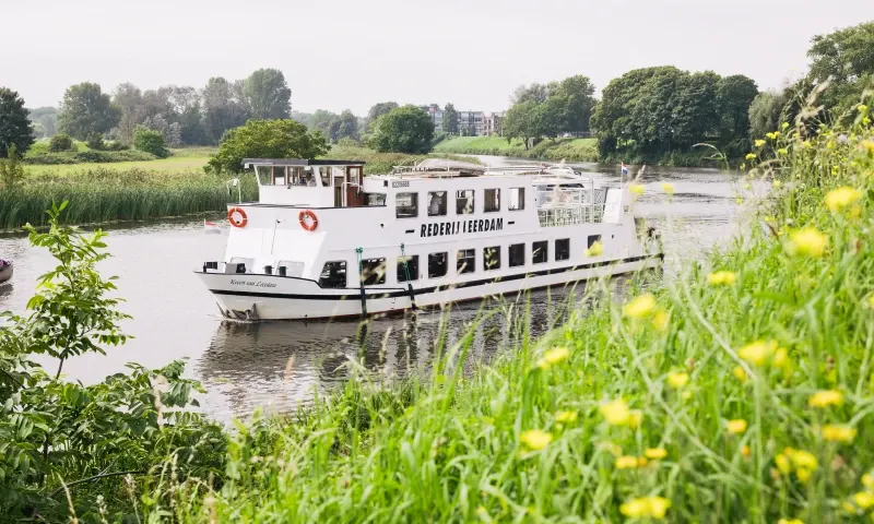 Het cruiseschip van Rederij Leerdam vaart over het water, op de voorgrond gele bloemen.