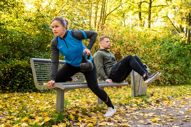 Een man en vrouw zijn samen aan het sporten in het bos. Ze doen verschillende oefenen op een bankje die in het bos staat.