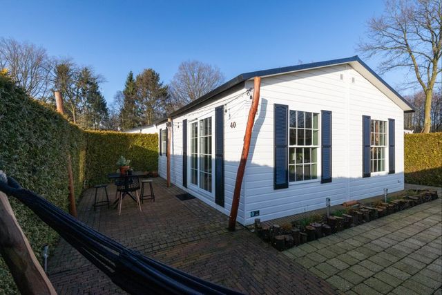 Een wit houten chalet met blauwe luiken, terras met op de voorgrond een hangmat, blauwe lucht.