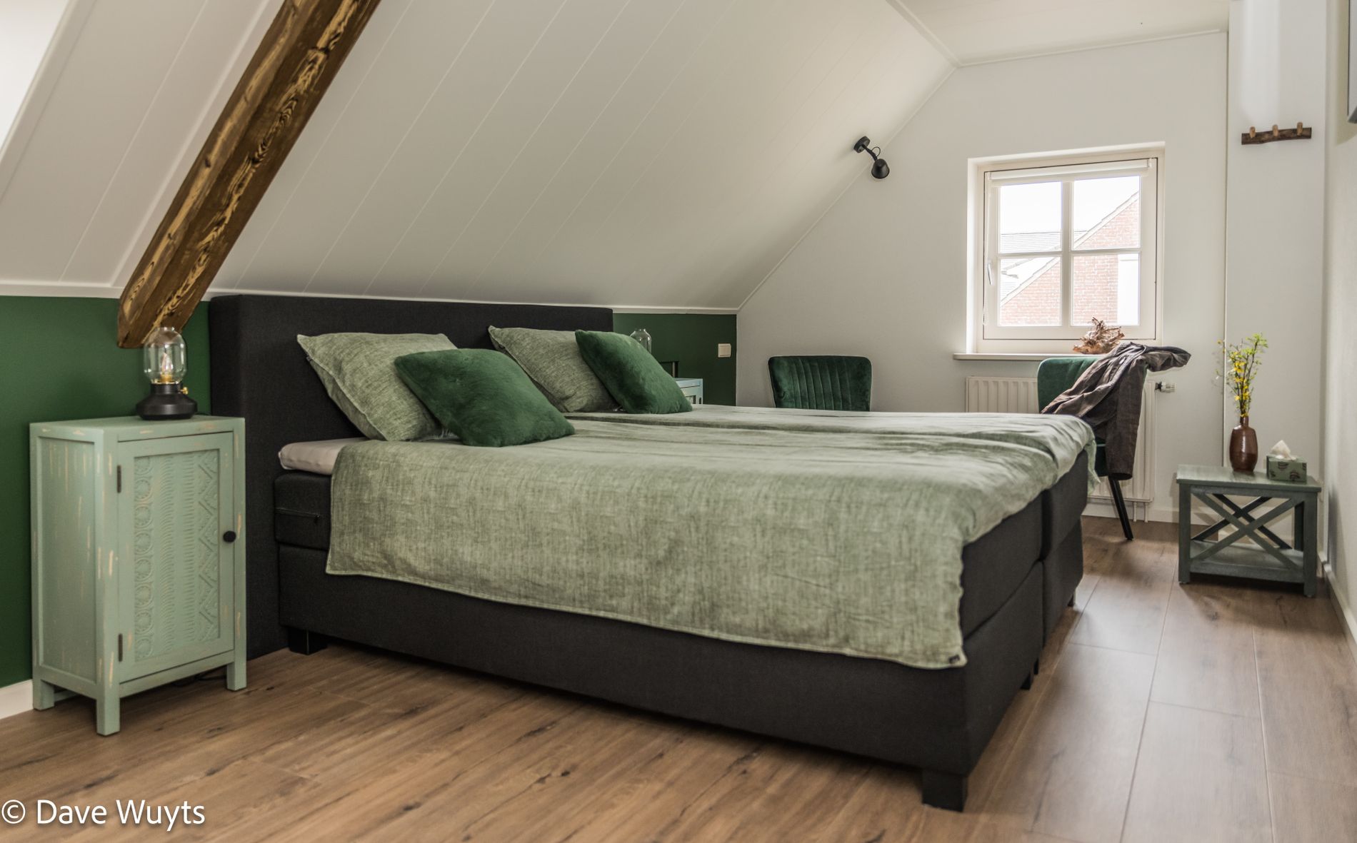 Slaapkamer met twee-persoons bed met groene accenten onder de schuine kap. Onder het raam in de gevel een zitje in groen.