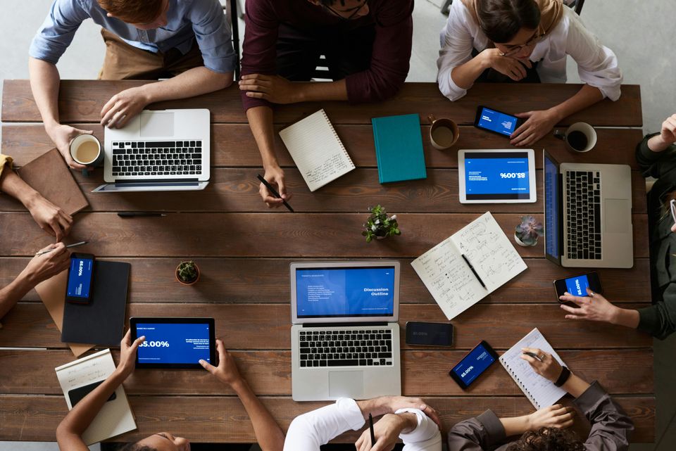 Tafel met 7 mensen die vergaderen, er staan diverse laptops en iPads op tafel.