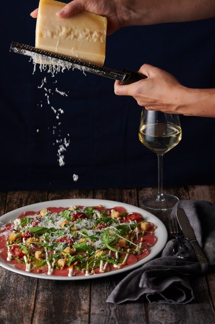 Hand raspt Parmezaanse kaas over carpaccio met rucola, pijnboompitten en croutons, geserveerd met een glas witte wijn bij Bistro de Bock in Woerden.