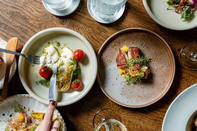 Bovenaanzicht van verfijnde gerechten op een houten tafel in restaurant Bistronoom, met onder andere burrata, gegrilde groente, cherry tomaatjes en een stukje langzaam gegaard vlees.