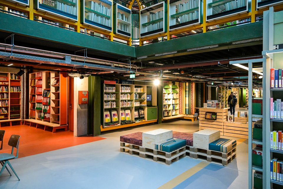 Overzicht van een moderne bibliotheek met open indeling: boekenkasten langs de wanden, zitblokken in het midden, een informatiebalie en een bezoeker die door de ruimte loopt.