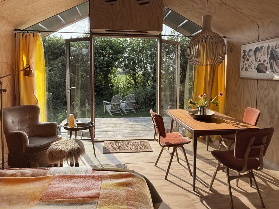 Interieur van een wikkelhuisje bij Proeftuin van Linschoten, met houten wanden, eettafel en zithoek, en openslaande deuren naar een terras met stoelen in het gras naast hoge struiken.