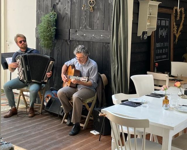 Twee mannen zitten op een stoel en spelen muziek waarvan er eentje de gitaar speelt en de andere accordeon.