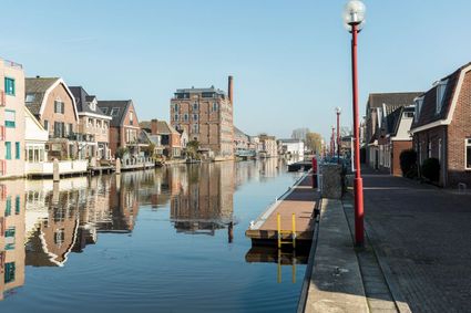 Rustige Nederlandse stadsgracht met historische huizen aan beide zijden, weerspiegelingen in het kalme water, een wandelkade met rode lantaarnpalen en een aanlegsteiger op de voorgrond onder een heldere blauwe lucht.