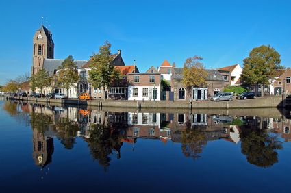 Historische gevels en de Grote Kerk van Oudewater langs een stille gracht, met spiegeling in het water en een heldere blauwe lucht.