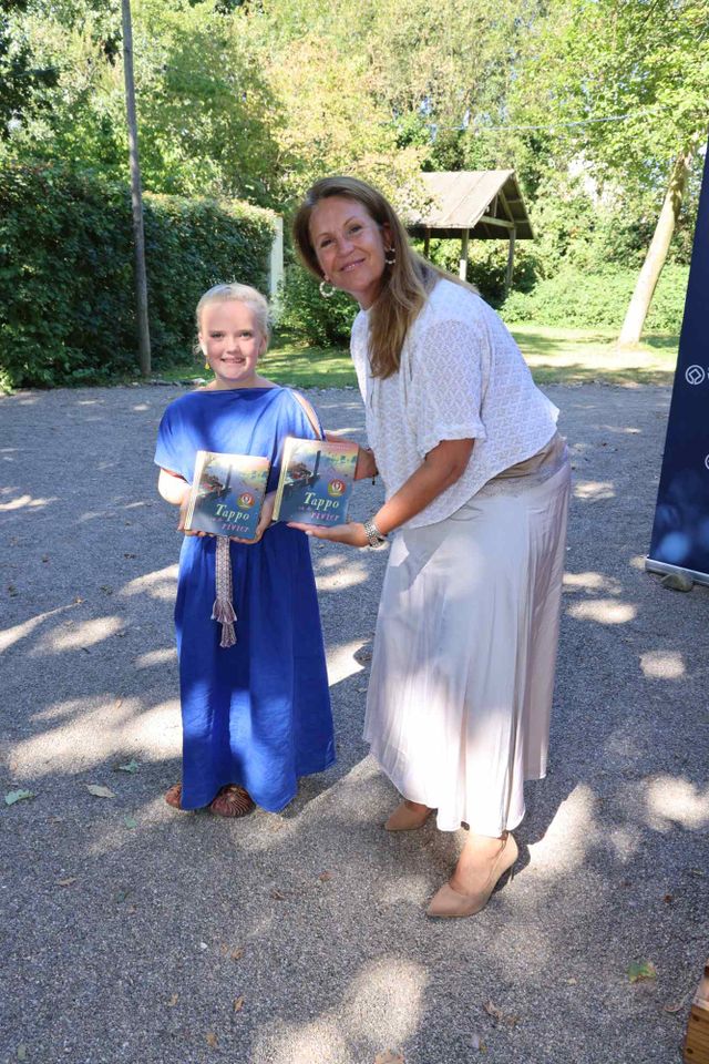 Een meisje in een blauwe romeinse jurk naast wethouder Chantal van den Berg van Alphen aan den Rijn, met het boekje Tappo in de hand.