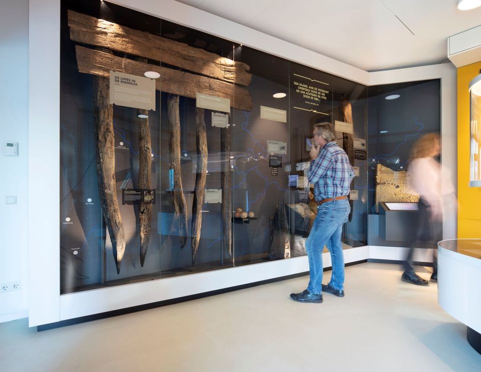 Bezoeker in de houtkamer van Museum Hoge Woerd. In de vitrine zijn diverse houten gepunte palen van de Limesweg te zien.