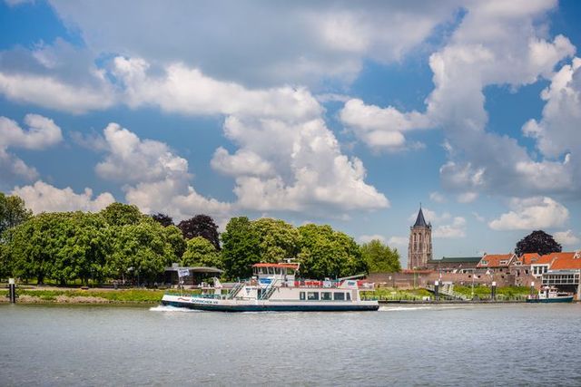 Veerboot vaart over de Merwede met uitzicht op de skyline van Gorinchem, inclusief stadsmuur, kerktoren en groene bomen onder een wolkenrijke lucht.