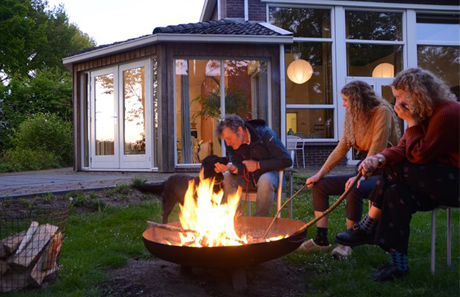 Drie mensen zitten rond een vuurschaal met kampvuur bij een groepsaccommodatie.