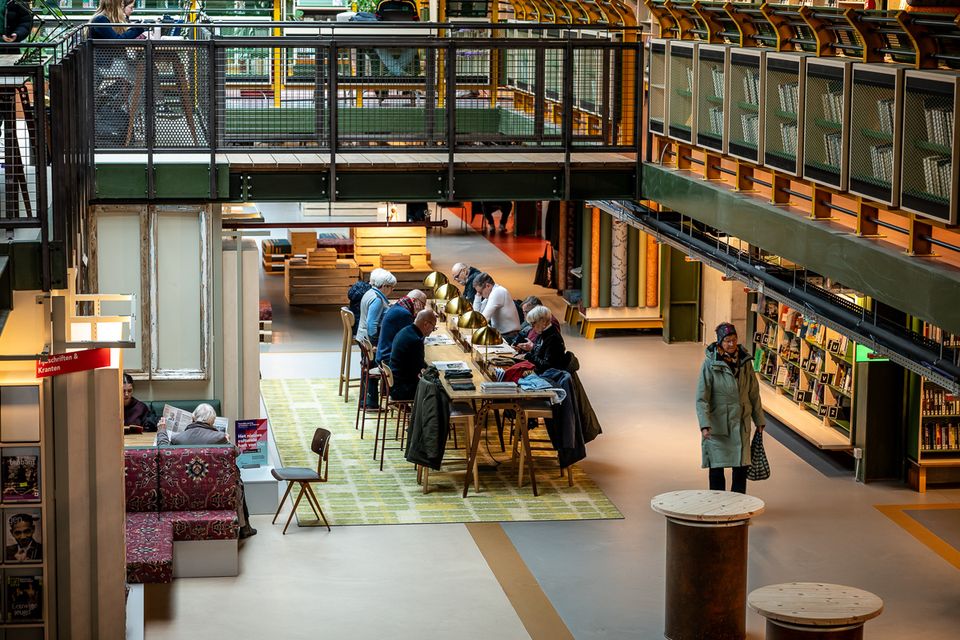 Bezoekers lezen en werken aan een lange leestafel in een open bibliotheek met meerdere verdiepingen, omringd door boekenkasten, looproutes en een industriële brugconstructie.
