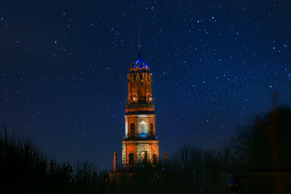 Een indrukwekkend nachtelijk tafereel: de verlichte kerktoren rijst op tegen een diepblauwe, met sterren bezaaide hemel. Het warme licht van de toren contrasteert prachtig met de stille duisternis eromheen, waardoor het monument een bijna tijdloze en serene uitstraling krijgt.
