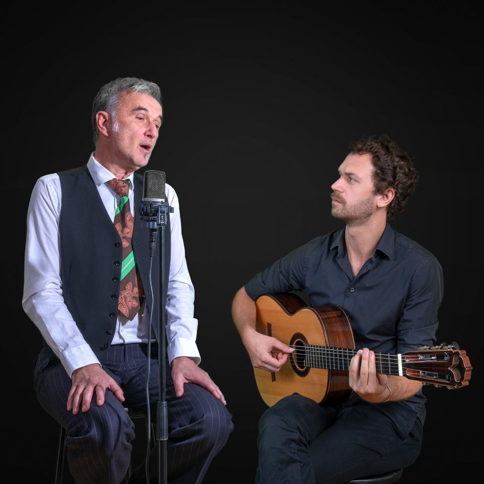 Foto van de zanger, zittend op een kruk achter een microfoon en de gitarist, zittend met zijn gitaar op schoot tegen een zwarte achtergrond.