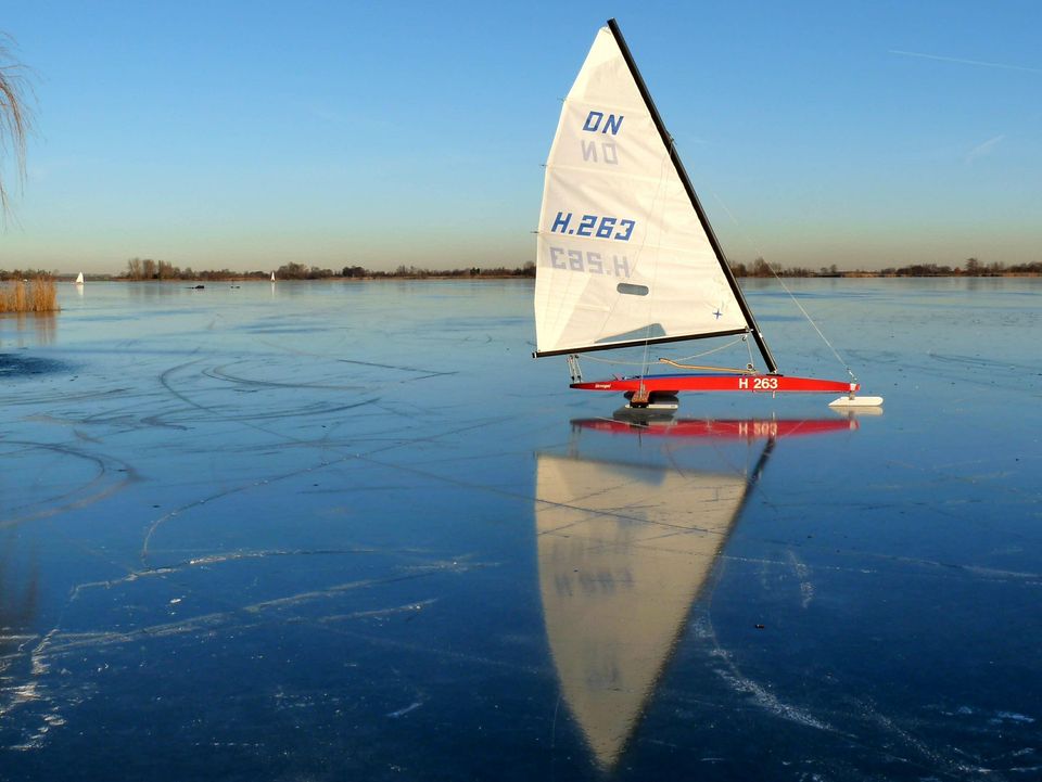 Een verstild winterlandschap op het ijs: een ijszeiler glijdt geruisloos over een spiegelgladde, bevroren plas, met het witte zeil scherp afgetekend tegen de heldere blauwe lucht. De perfecte weerspiegeling in het ijs en de uitgestrekte ruimte geven het beeld een gevoel van vrijheid en winterse stilte.