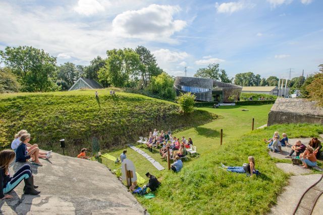 Een glooiende omgeving met veel gras waarin een modern paviljoen staan. Overal zitten mensen.