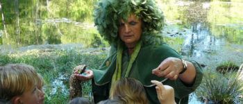 Foto van een vrouw met een groene verenpruik voor een vijver met kinderen om haar heen in een bosrijke omgeving.