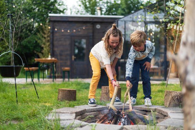 Broer en zus leggen een tak op het kampvuur bij hun vakantiehuisje.