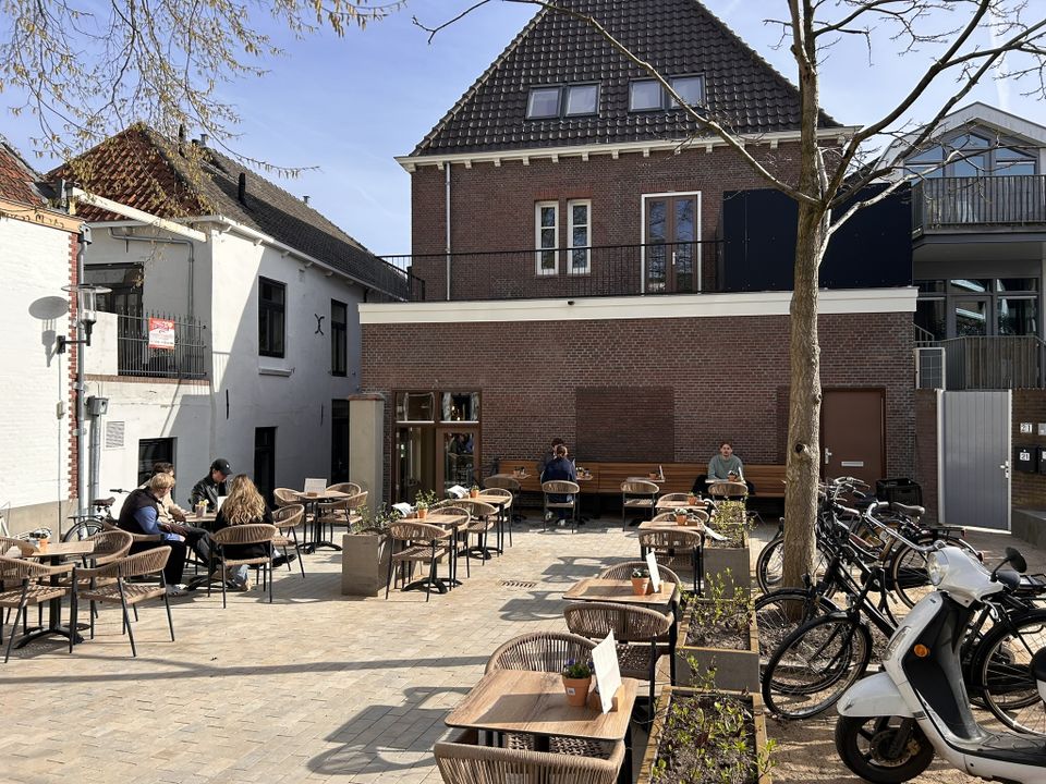 Stadsterras Barista Café Woerden in Woerden, Groene Hart, met gasten op het zonnige terras, houten tafels en stoelen, fietsen geparkeerd langs de rand en moderne bebouwing rondom het plein.
