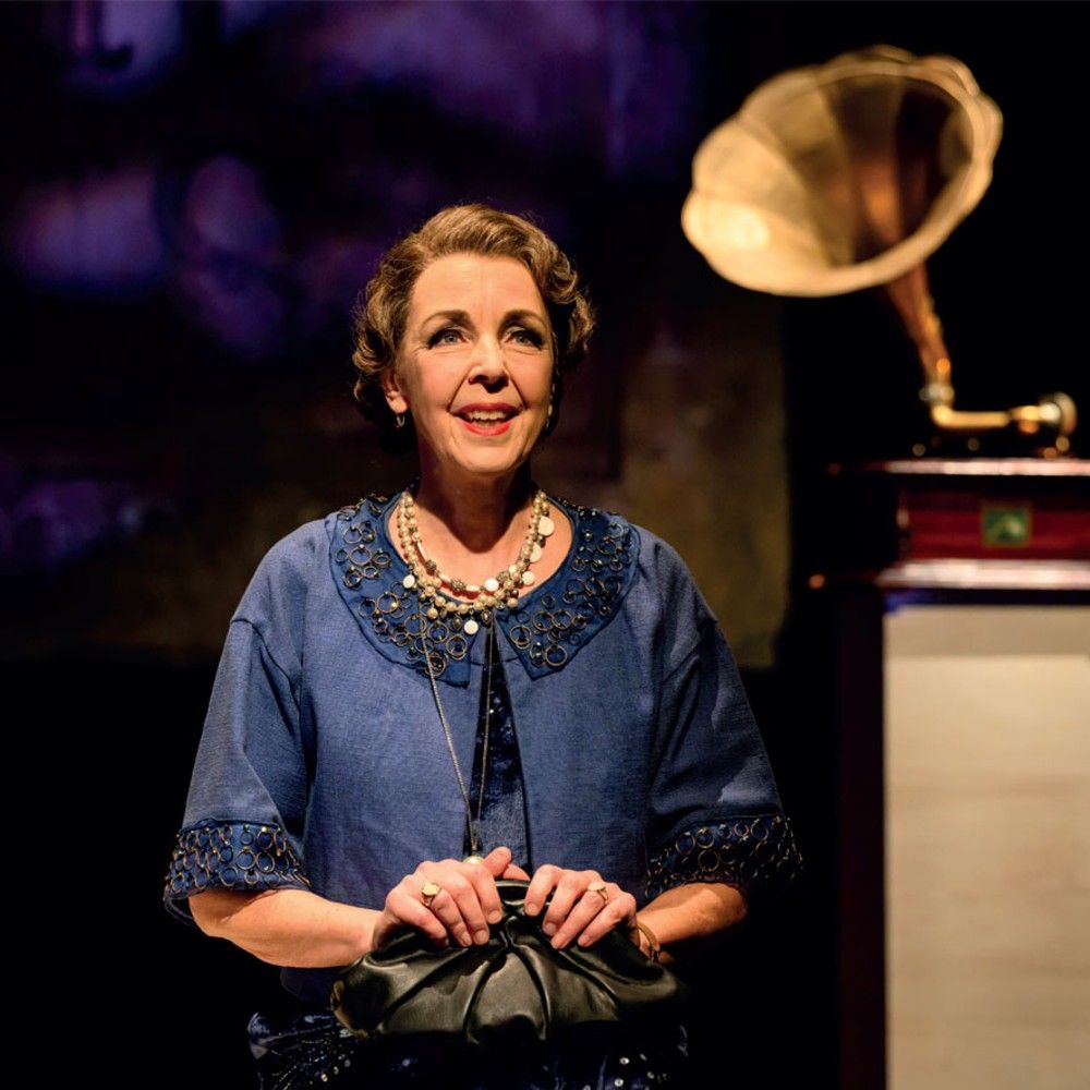 Foto van een dame in een blauwe jurk met juwelen en een zwart tasje op haar schoot, zittend voor een oude grammofoon.