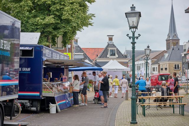 Een vrouw loopt met haar fiets langs de viskraam op de weekmarkt in Lemmer. Er staat een rij bij de viskraam en er staan lange houten tafels en bankjes om op te zitten. Op de achtergrond staat de kerktoren.