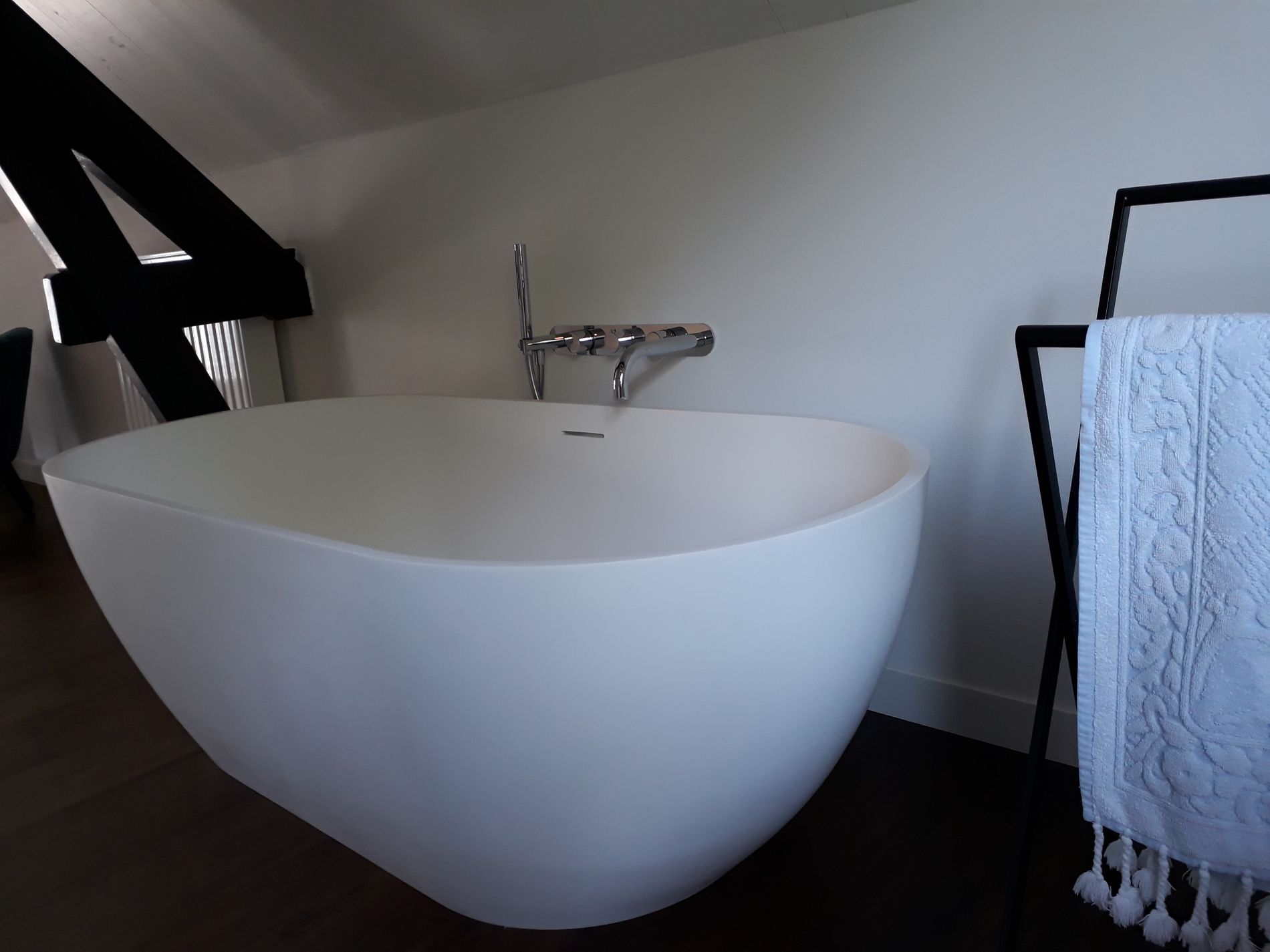 Detailfoto van een wit ligbad onder schuine kap met donkere spanten in het zicht. Rechts staat een donker handdoekrekje met witte handdoek.