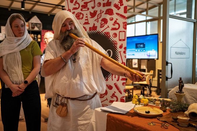 Een man blaast op een hoorn, bezoekers kijken toe.