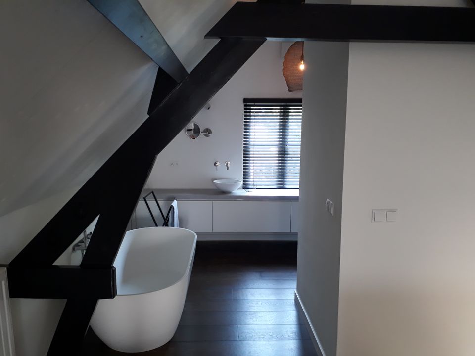 Een wit ligbad onder schuine kap met donkere spanten in het zicht. Een donker handdoekrekje met witte handdoek voor een lage kastenwand met wastafel onder het raam.