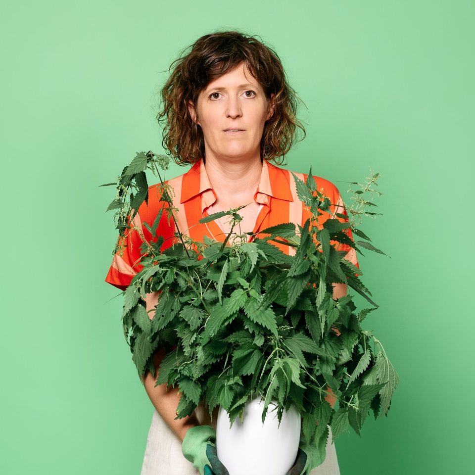 Foto van Janneke met een oranje bloesje aan tegen een groene achtergrond. Ze houdt een witte vaas met een grote bos brandnetels vast.