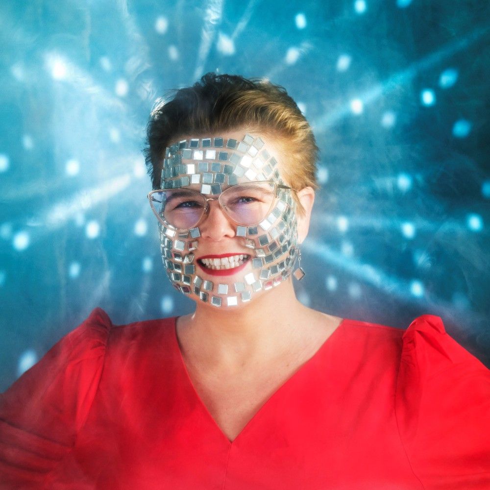 Foto van Marjolein in een rode top tegen een lichtblauwe achtergrond. Ze heeft glitters op haar gezicht geplakt die reflecteren in de achtergrond.