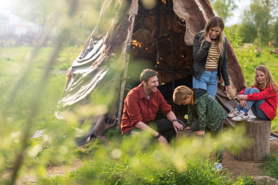 Een gezin met twee kinderen zit voor een hut in het hunebedcentrum.