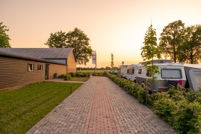 Foto van een klinkerpad met aan de linkerkant een bijgebouw en aan de rechterkant diverse caravans in de avondzon.