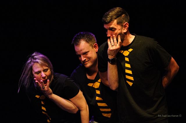 Acteurs van Allure geel in actie. Een vrouw in het midden knijpt in de de wangen met haar hand van de mannen aan beide zijden.