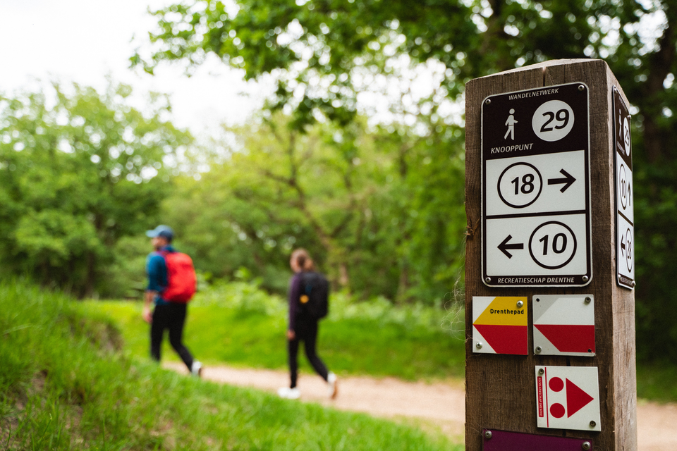 Een paaltje met routebordjes en twee wandelende mensen verderop