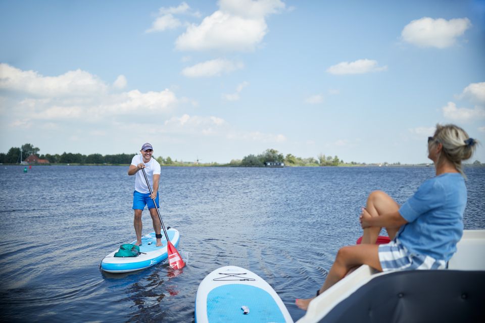 En man staat op een SUP-board op het water en lacht naar een vrouw die op een boot zit.