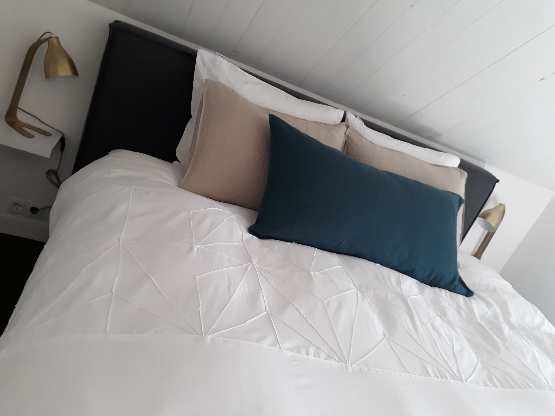 Detailfoto van een twee-persoonsbed, wit beddengoed en zand- en donkerblauwkleurige kussens, onder een schuine kap.