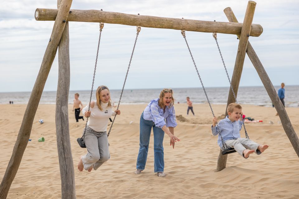 Twee kinderen schommelen met hun moeder op het strand van Katwijk aan Zee, met de zee op de achtergrond.