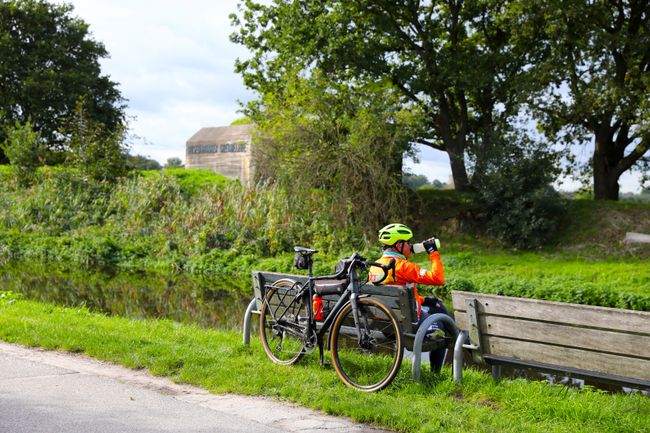 Wielrenner zit op een bankje met zijn fiets achter zich. Voor hem zie je tussen het groen een bunker van de Grebbelinie en de rivier.