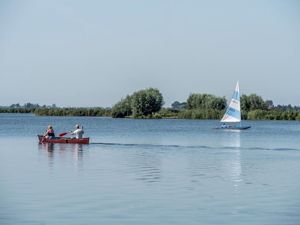 In de verte varen 2 mensen in een kano met een zeilbootje op de achtergrond.