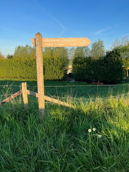 Houten wegwijzer “Hollandse IJsselpad” in groen weiland bij avondlicht, met pijl naar ’t Klaphek → Gouda (37 km) onder heldere blauwe lucht.