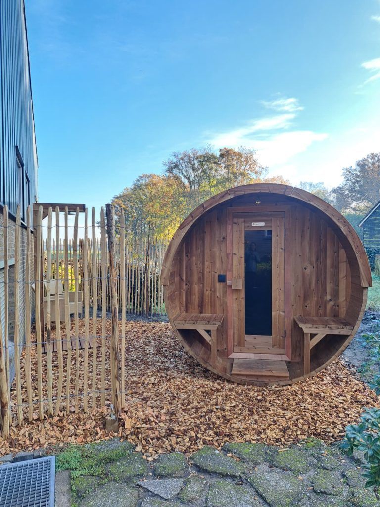 Ronde houten barrelsauna met glazen deur en zitbankjes staat op houtsnippers in een tuin naast een gebouw en houten hekwerk.