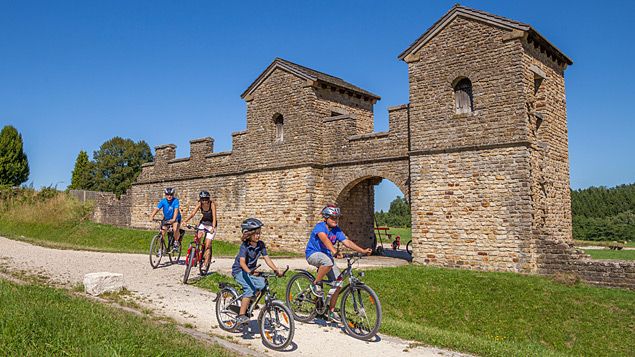 Fietsers met helmen voor een herbouwde Romeinse poort in een groen landschap