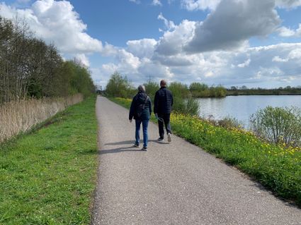 Wandelroute in het Groene Hart, waar twee wandelaars over een verhard pad langs water, rietkragen en groen natuurgebied lopen onder een wolkenrijke voorjaarslucht.