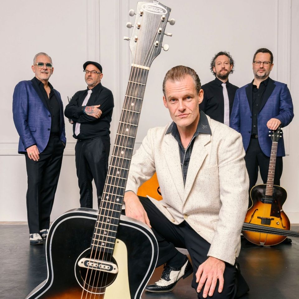 Foto van de band, 5 heren met hun muziekinstrumenten in blauw-zwart-wit gekleed, voor een lichte achtergrond.