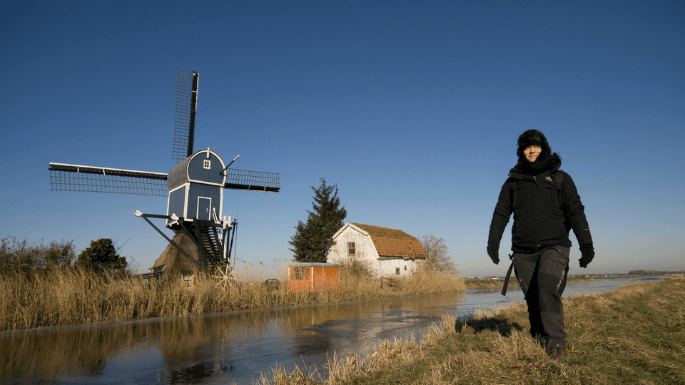 Wandelaar tijdens de winter langs het water met op de achtergrond een molen.