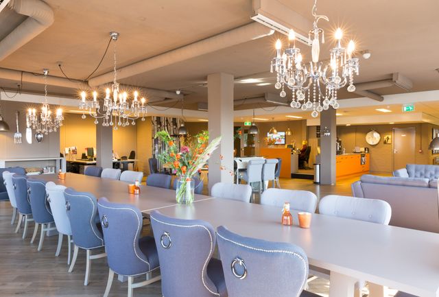 Stijlvolle vergaderruimte bij Hofclub Werken & Vergaderen met lange tafel, blauwe stoelen, kroonluchters en bloemen op tafel – warme, moderne loungeomgeving in Woerden.