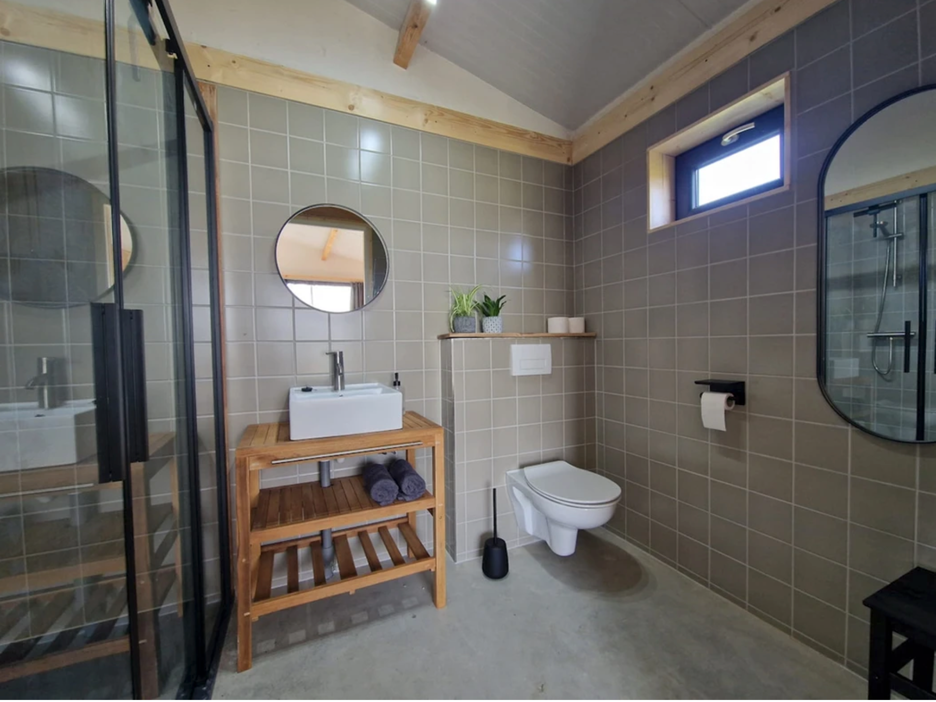 Douche en wc met grijze tegels, zwarte en houten accessoires.