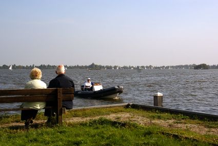 Een ouder echtpaar zit op een bankje met uitzicht over de plassen. Op de plassen vaart een sloep dicht bij de kant