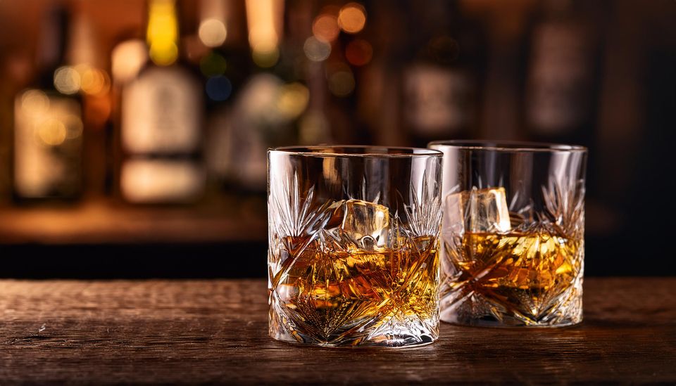 Twee lage glazen met whisky en ijsblokjes staan op een houten bar. Op de achtergrond zijn flessen drank zichtbaar, onscherp weergegeven, wat zorgt voor een warme en sfeervolle setting.